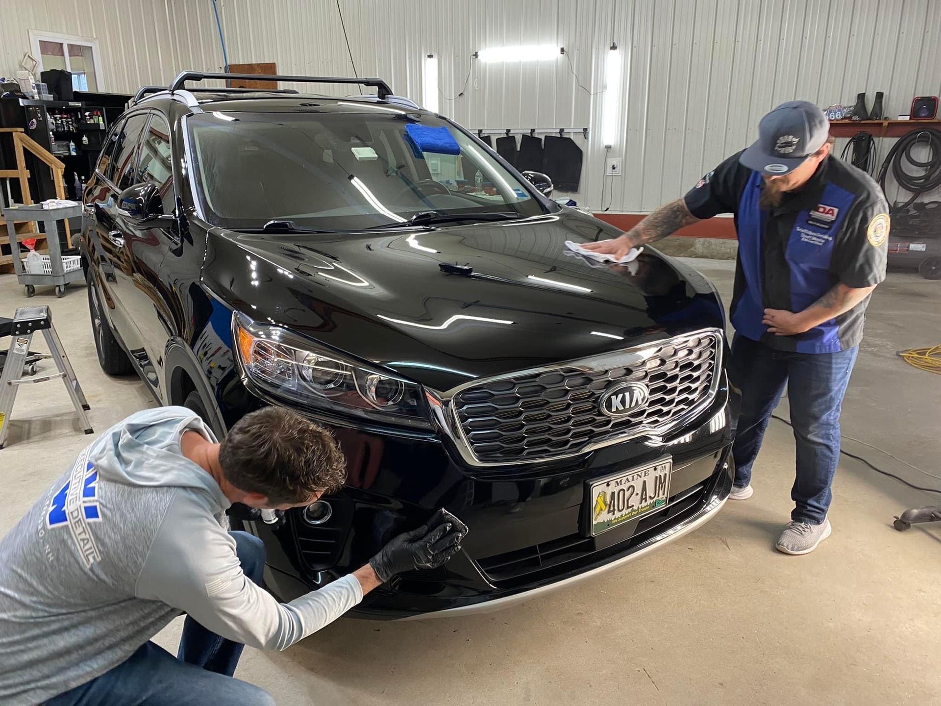 Two men detailing a black Kia SUV in a garage; one is wiping the hood, the other the bumper.