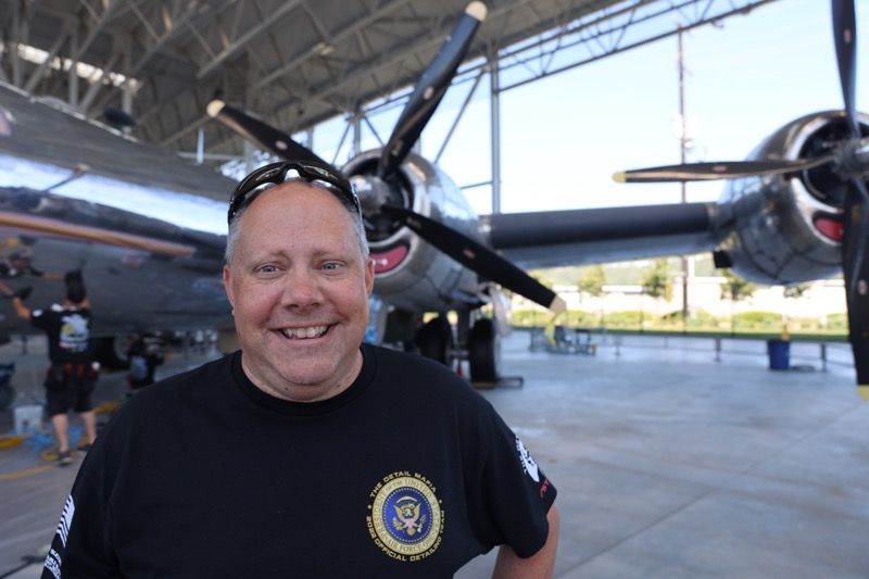 Man in black shirt smiling, posing in front of a vintage airplane with propellers. Outdoors, sunny day.