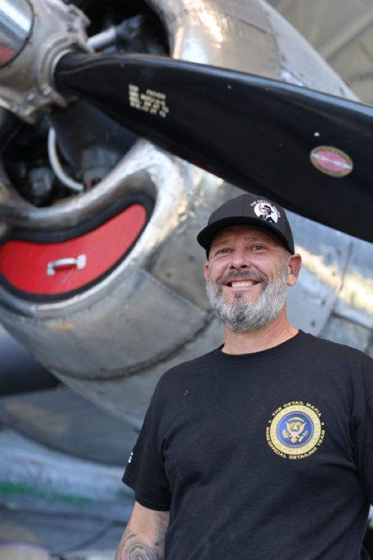Man with graying beard smiles in front of a plane propeller. He wears a black cap and t-shirt.