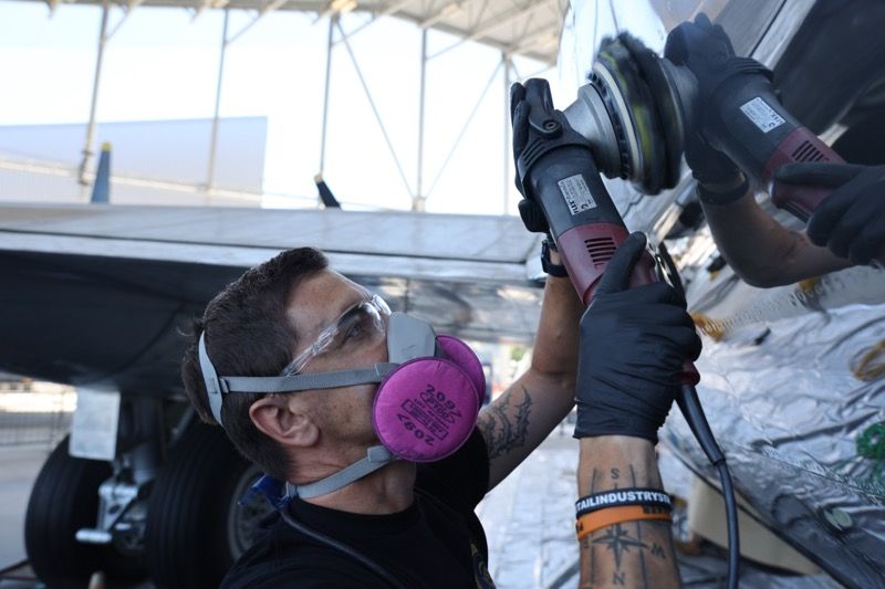 Man wearing safety gear sanding a silver airplane wing.