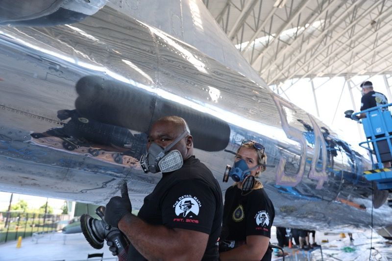 Two people in masks polishing a silver aircraft in a hangar.