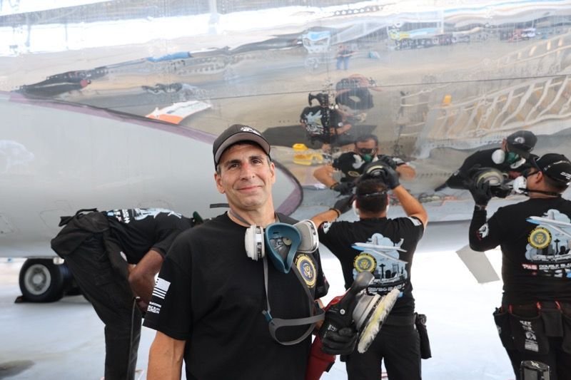 Man in black shirt smiles in front of a polished aircraft. Workers in similar shirts repair the plane.