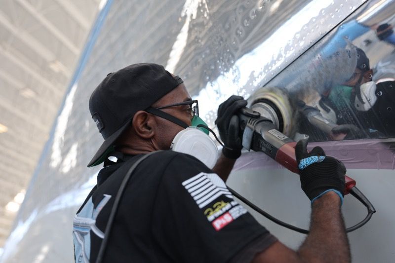 Man polishes a vehicle's side with a power tool, wearing a mask, gloves, and a cap, outdoors.