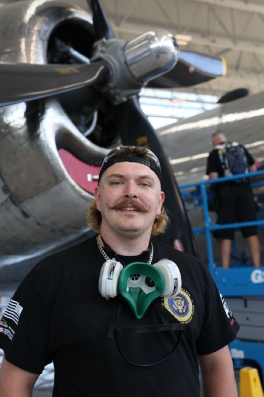 Man with a mustache wearing a respirator in front of a vintage airplane.