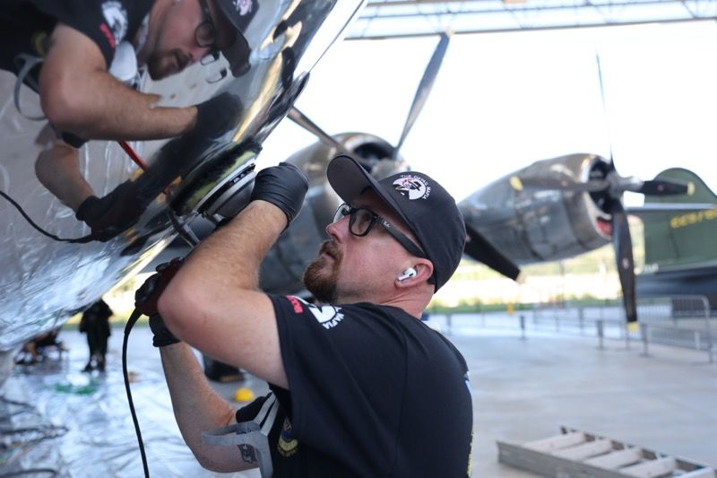 Two men working on the wing of a vintage airplane outdoors. One is using a tool.