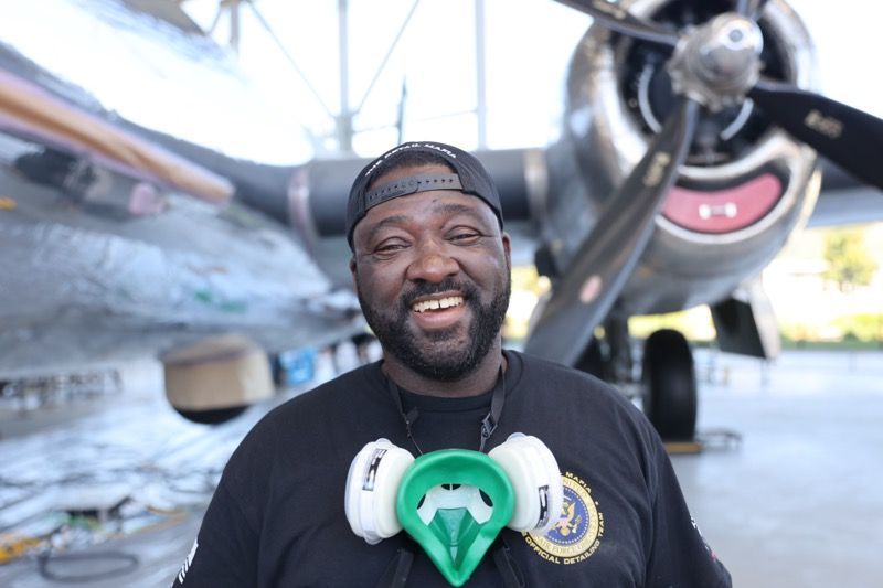 Man with beard smiles in front of a vintage airplane, wearing a respirator around his neck.
