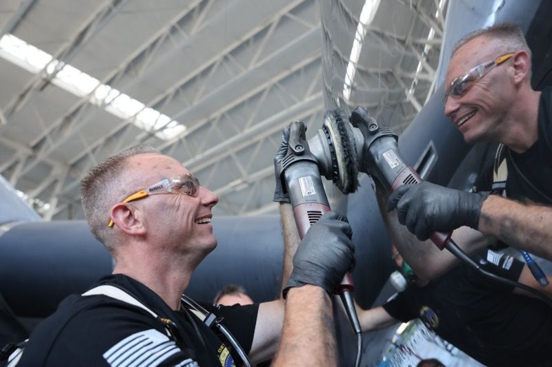 Two men in a hangar, smiling, use power sanders on an aircraft. They wear safety glasses and gloves.