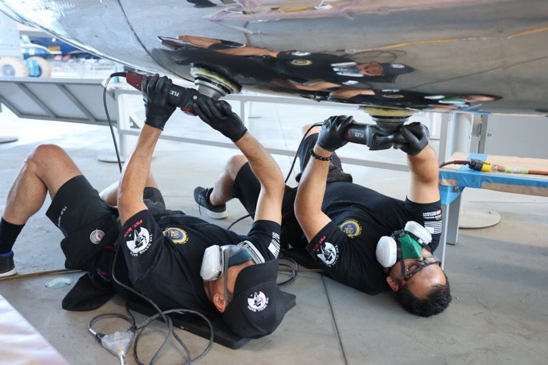 Two men polishing airplane fuselage lying on their backs, wearing safety gear.