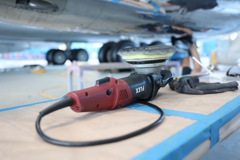 Red and black power polisher on a workbench under an aircraft, used for surface work.