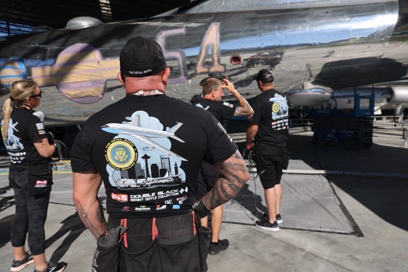 Crew members in black shirts with logos work on a silver airplane, likely in a hangar.