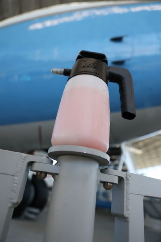 Pink liquid dispenser on a gray post, near an airplane.