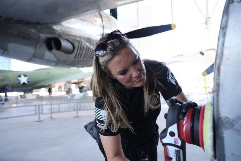 Woman polishing an airplane propeller inside a hangar; wearing sunglasses and a black shirt.