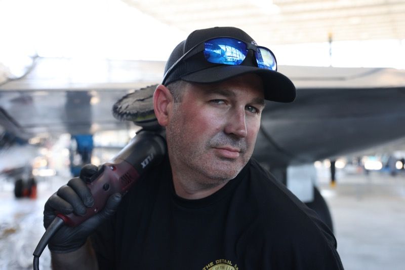 Man holding a power tool, wearing black cap and sunglasses, near an airplane.