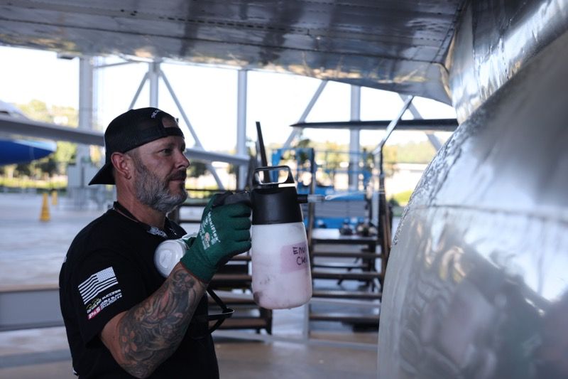 Man sprays aircraft with cleaning solution in an outdoor hangar, wearing a mask and gloves.
