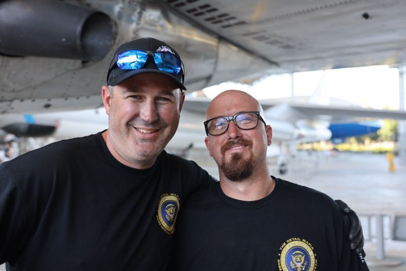 Two men smiling in front of an airplane. Both wearing black shirts, one with a hat, the other with glasses and a beard.