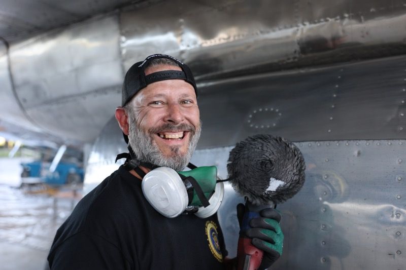 Man smiles, holds polisher near airplane fuselage, wearing cap, safety gear, and gloves.