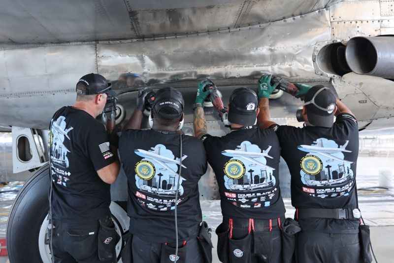 Four people in matching shirts work on the underside of an aircraft, using tools.
