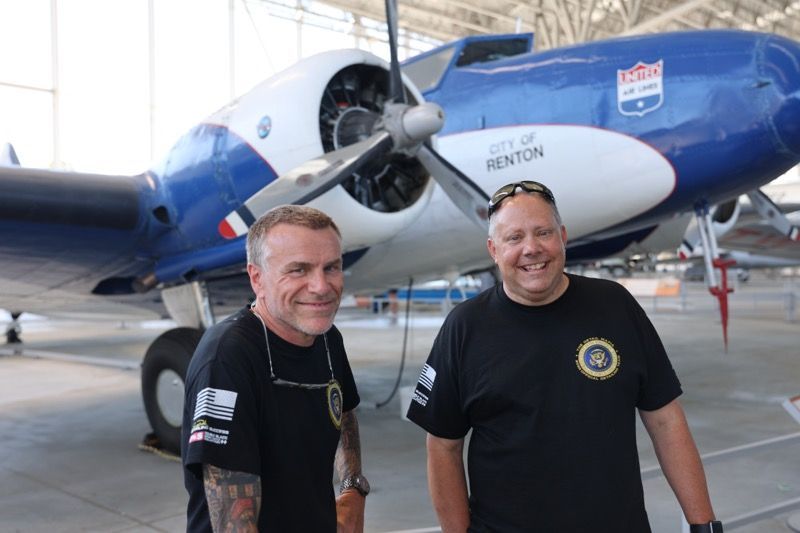 Two men stand smiling in front of a blue and white vintage airplane labeled 