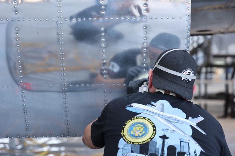 Man working on an airplane, using a tool, reflecting on the plane's metal side. Wearing a black shirt with airplane design.