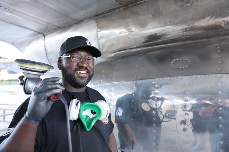 Man in black clothes and cap smiles, holding a sprayer near the silver airplane fuselage.