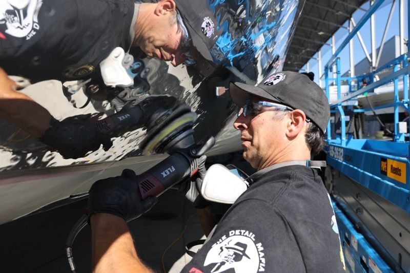 Two men polishing the hull of a boat with power tools. Outdoors on a lift. One is wearing a black cap and sunglasses.
