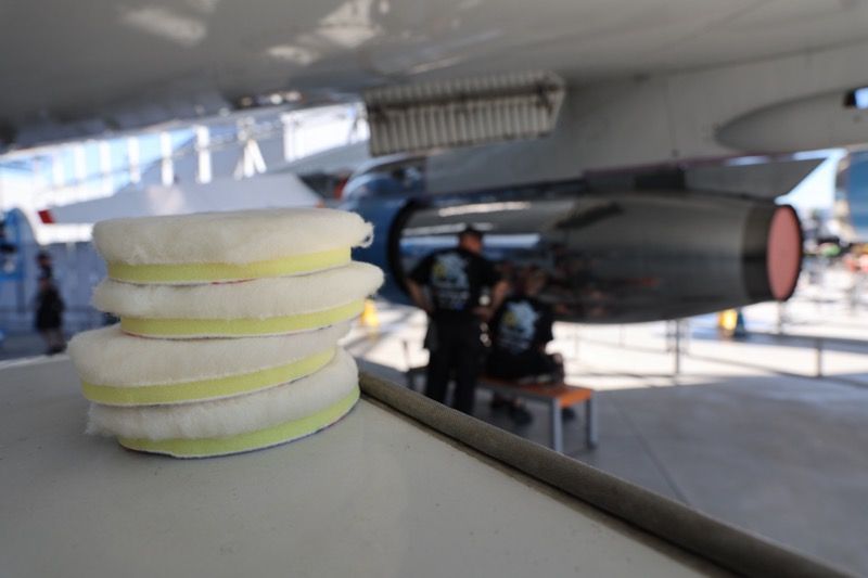 Stack of white and yellow polishing pads with airplane engine in background; two men.