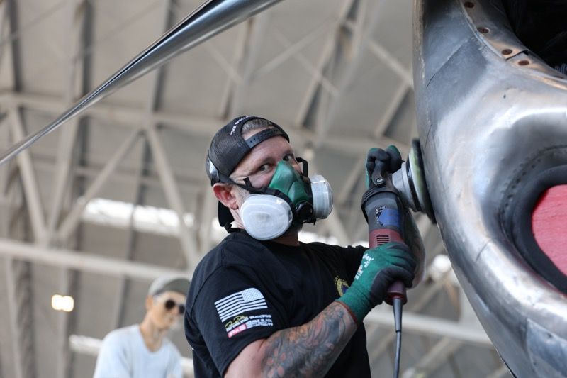 Man polishes aircraft propeller with power tool; wearing respirator and gloves in hangar.
