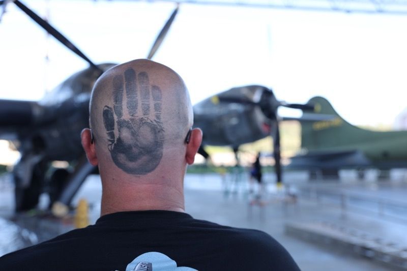 Bald man with a handprint tattoo on the back of his head stands in front of an airplane.