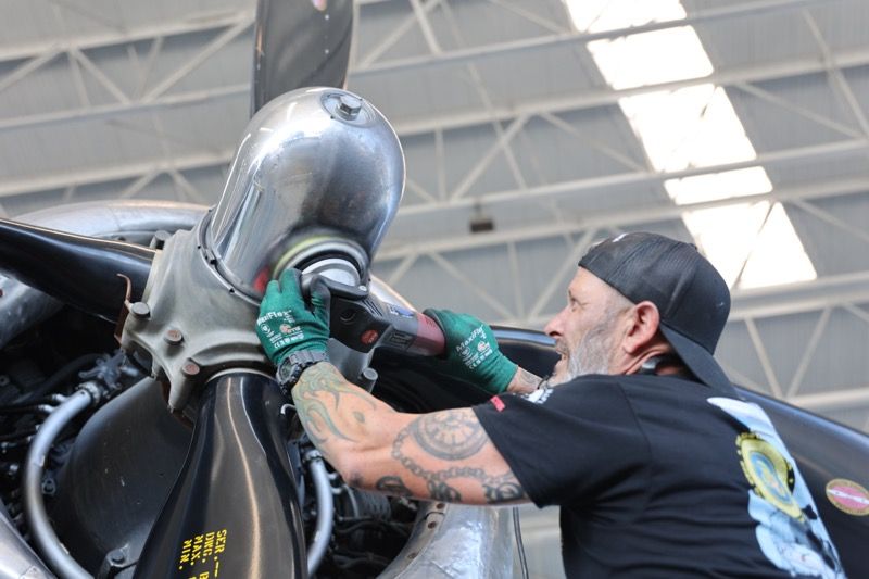 Man with tattoos works on an airplane propeller in a hangar, wearing gloves and a hat.