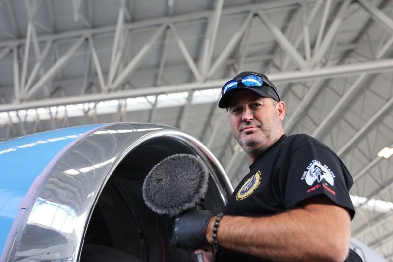Man polishing a blue aircraft wing, wearing black gloves and a cap, inside a hangar.