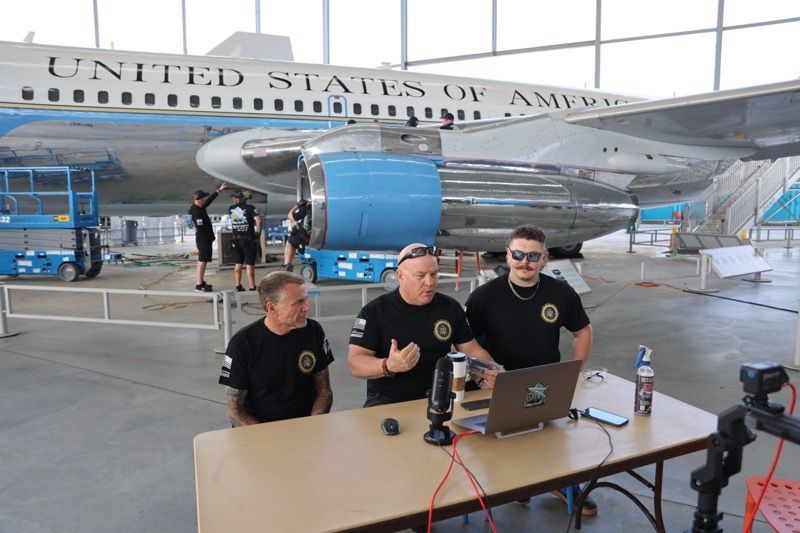 Three men in black shirts at a table with a laptop, microphone, and Air Force One in background.