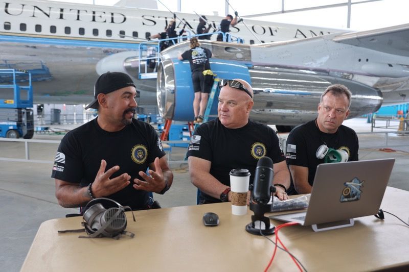 Three men at a table, recording a podcast with a plane in the background.