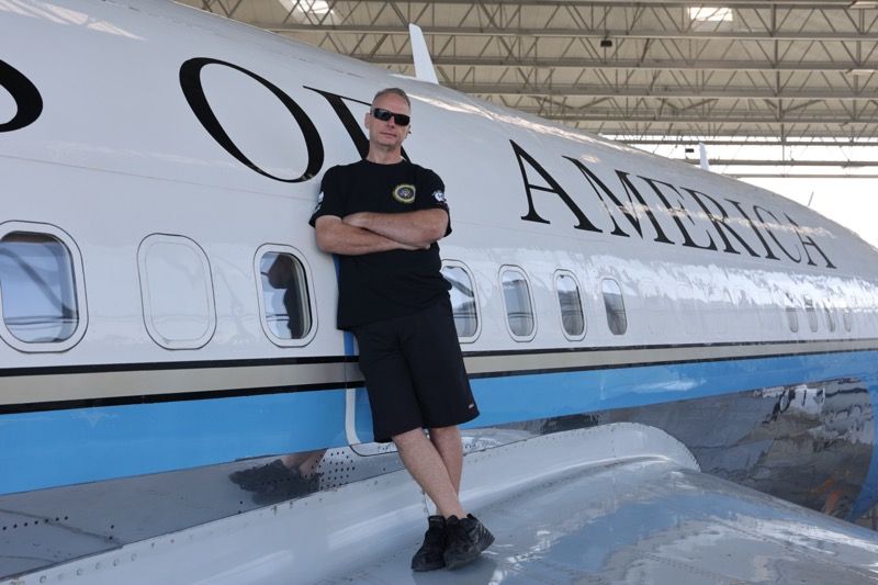 Man in sunglasses leans against Air Force One with arms crossed.