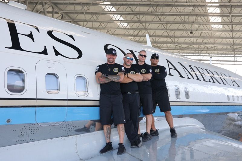 Four men in black clothing pose in front of a white and blue airplane, 