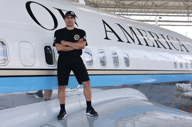 Man in black clothing stands with arms crossed on the wing of a plane labeled 
