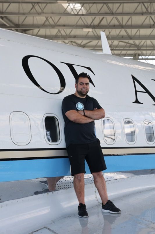 Man with arms crossed, smiling, stands by a white airplane with blue trim.
