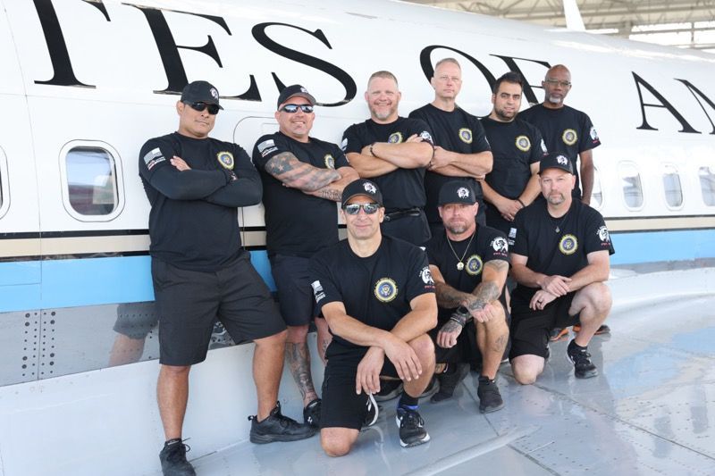 Group of men in black shirts pose in front of a plane that says “UNITED STATES OF AMERICA.”