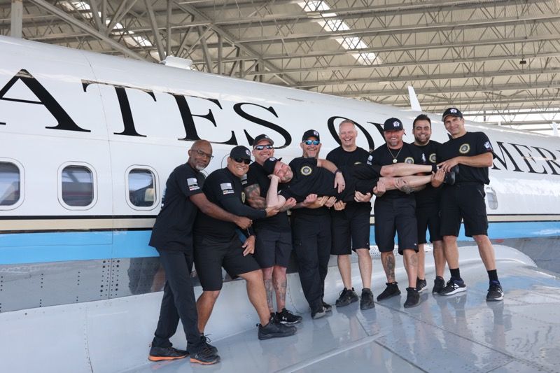 Group of men in black standing on plane wing, smiling. 