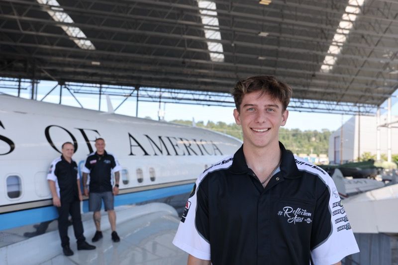 Smiling man in front of Air Force One with two other men, in a hangar.