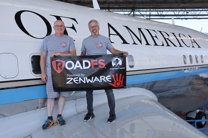 Two men pose with a banner in front of a plane labeled 