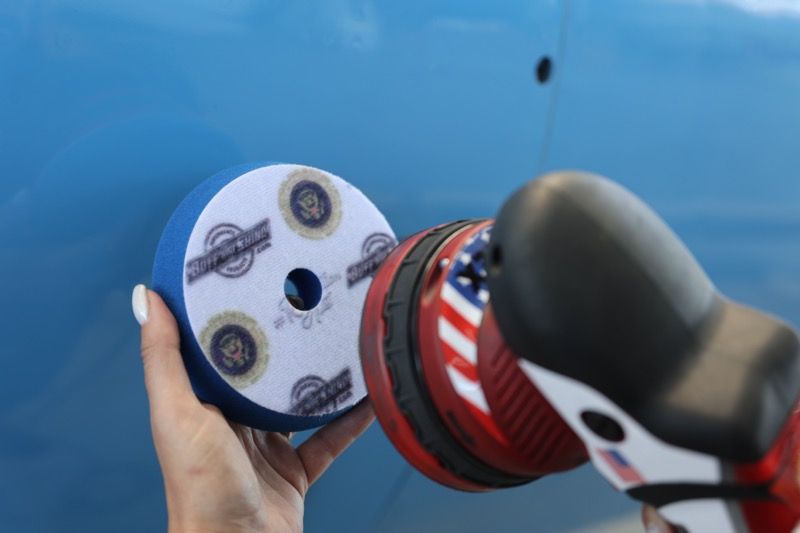 A woman holds a blue polishing pad near a red and white polisher on a blue car surface.
