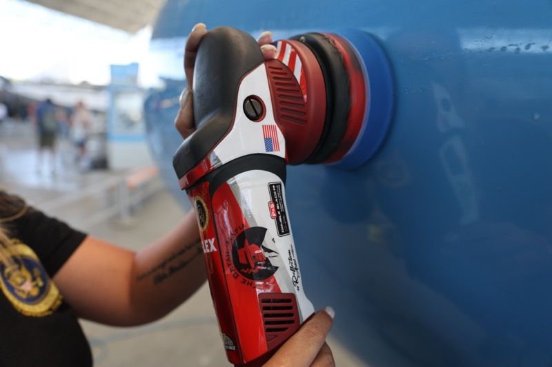 Person using a red and white polisher on a blue car's side; outdoors.