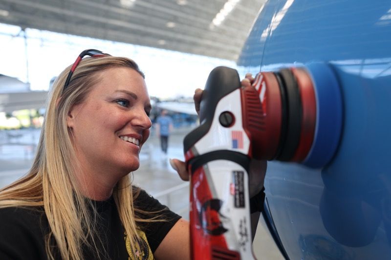 Woman polishes blue airplane with a power tool in a hangar, smiling.