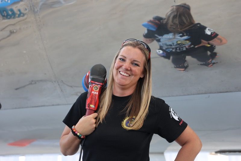 Woman with long blonde hair holding a red tool, smiling in front of an airplane wing; child reflected on wing.