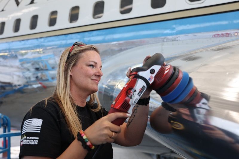 Woman using a power tool on an airplane wing in a hangar. She wears a black shirt and smiles.