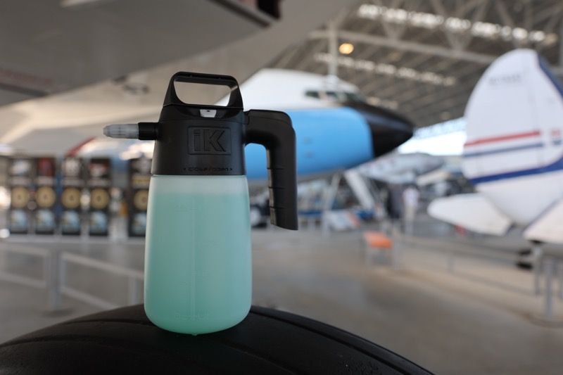 Foam sprayer with blue-green liquid, black handle, in a hangar with airplanes.