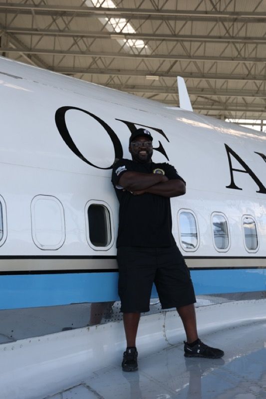 Man in black clothing stands smiling with arms crossed in front of a white airplane with the words 