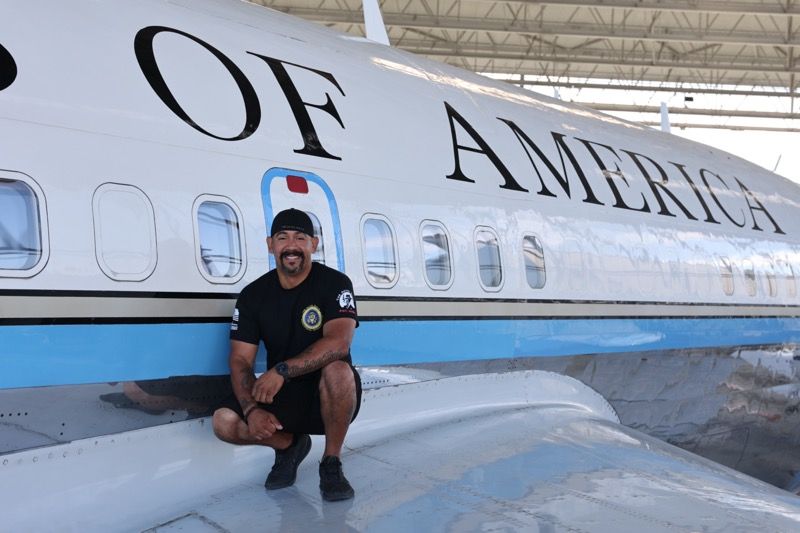 Man squats on airplane wing next to Air Force One; blue and white paint.