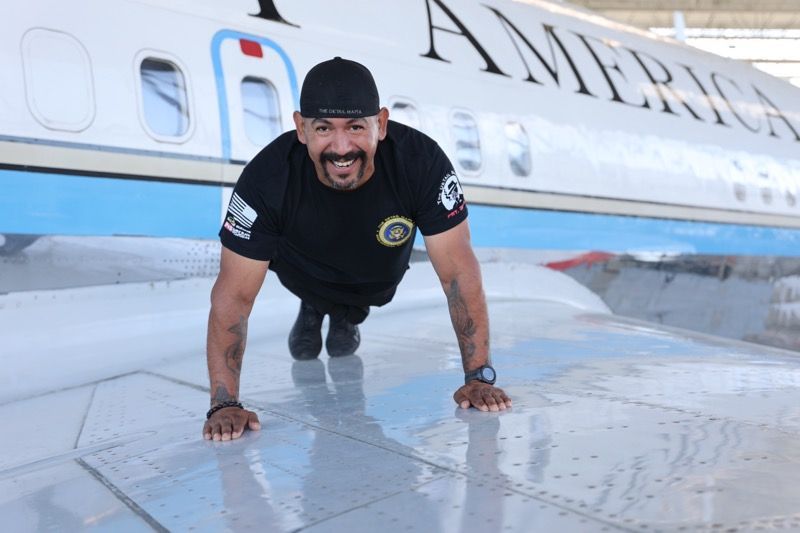 Man in black shirt doing a push-up on an airplane wing, smiling. 