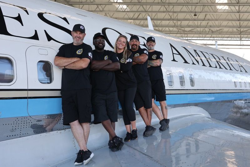 Group of five people in black shirts pose in front of an airplane; they're smiling with arms crossed.
